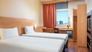 Habitación de hotel con dos camas, un escritorio, una silla, un televisor y ventana con vistas a edificios.