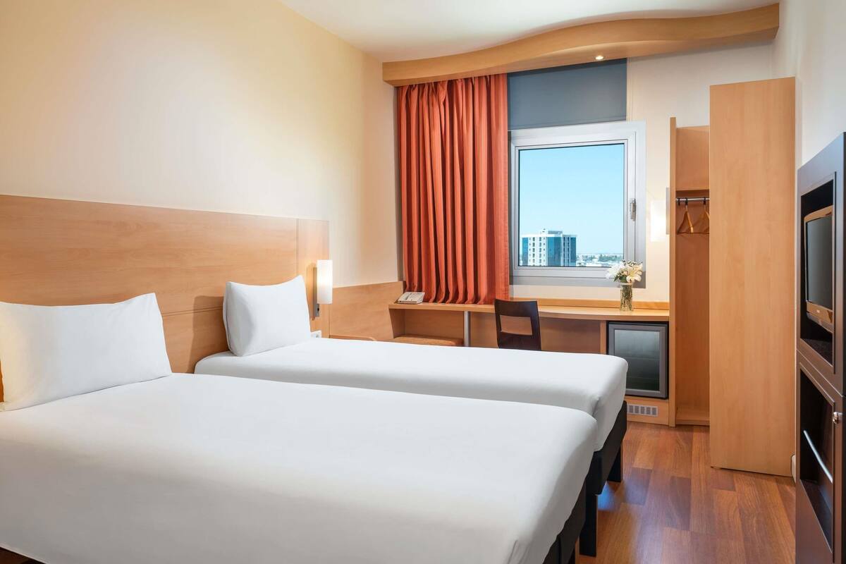 A hotel room with two beds, a desk, a chair, a TV, and a window with a view of buildings.