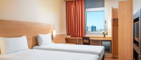 A hotel room with two beds, a desk, a chair, a TV, and a window with a view of buildings.