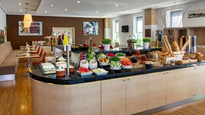 Un moderno vestíbulo de hotel con una zona de buffet central que ofrece una variedad de comidas y bebidas, un área de descanso con sillas rojas y una exhibición de arte contemporáneo en las paredes.