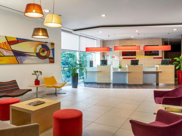 A modern hotel lobby with seating areas, a reception desk, and a large window providing a view of the outdoors.