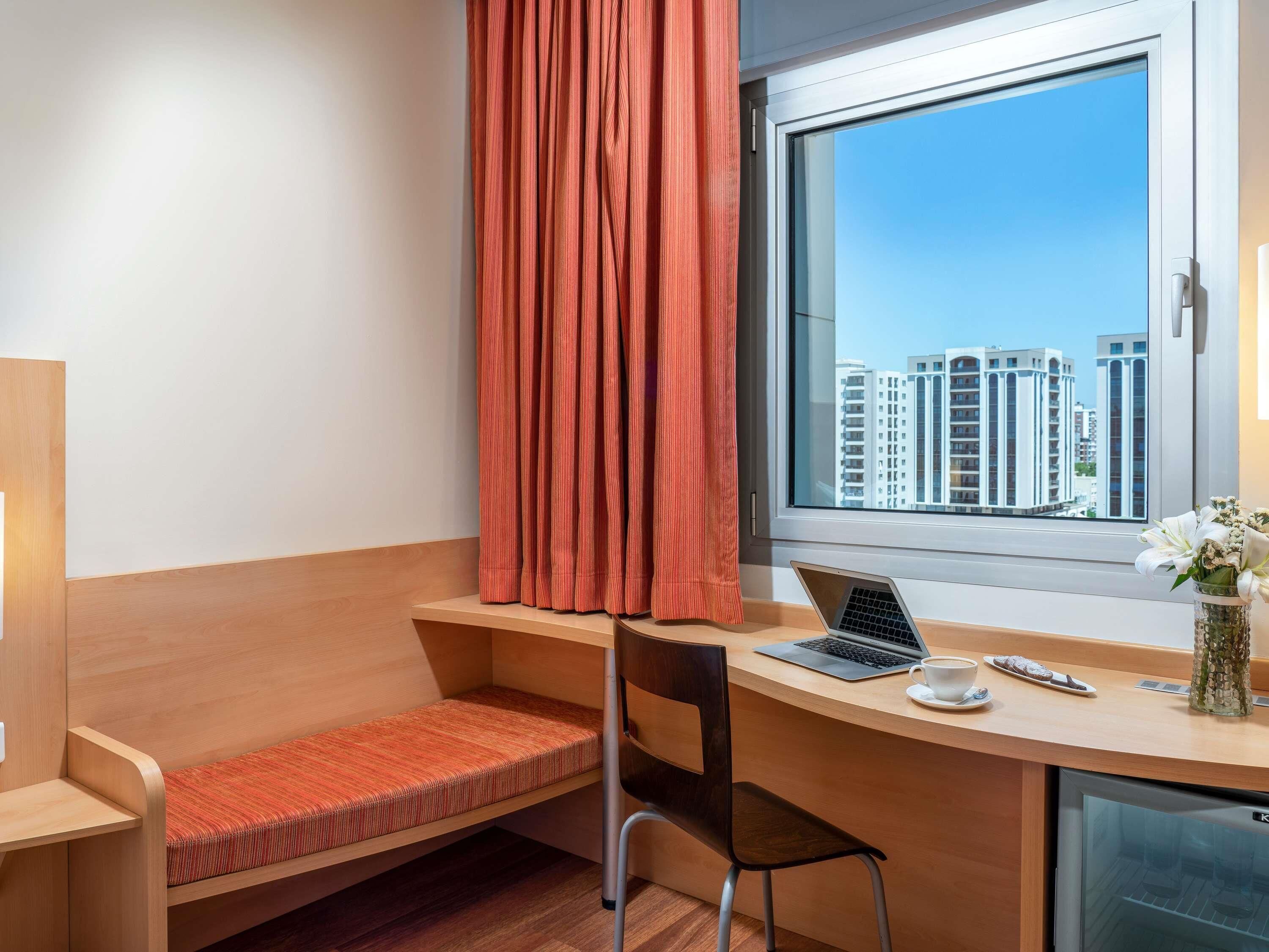 Habitación de hotel con escritorio, silla, ordenador portátil, taza de café y vistas a edificios.