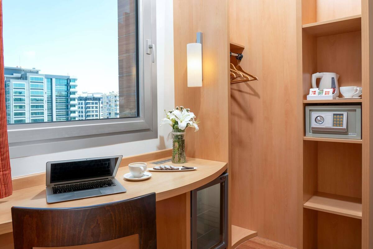 A hotel room with a wooden desk, a laptop, a cup of coffee, a vase with flowers, a safe, and a view of the cityscape through a window.