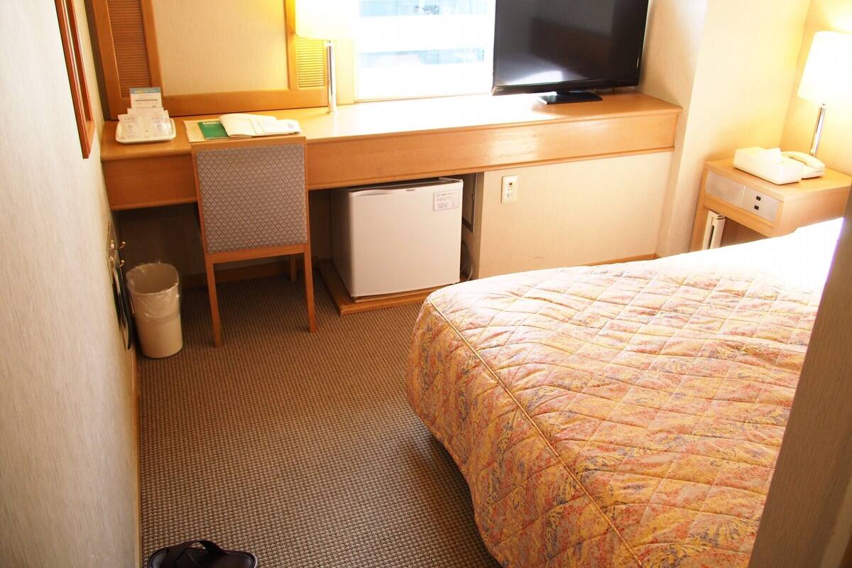 Une chambre d’hôtel comprenant un lit, un bureau avec une télévision, un petit réfrigérateur, une chaise et une poubelle.