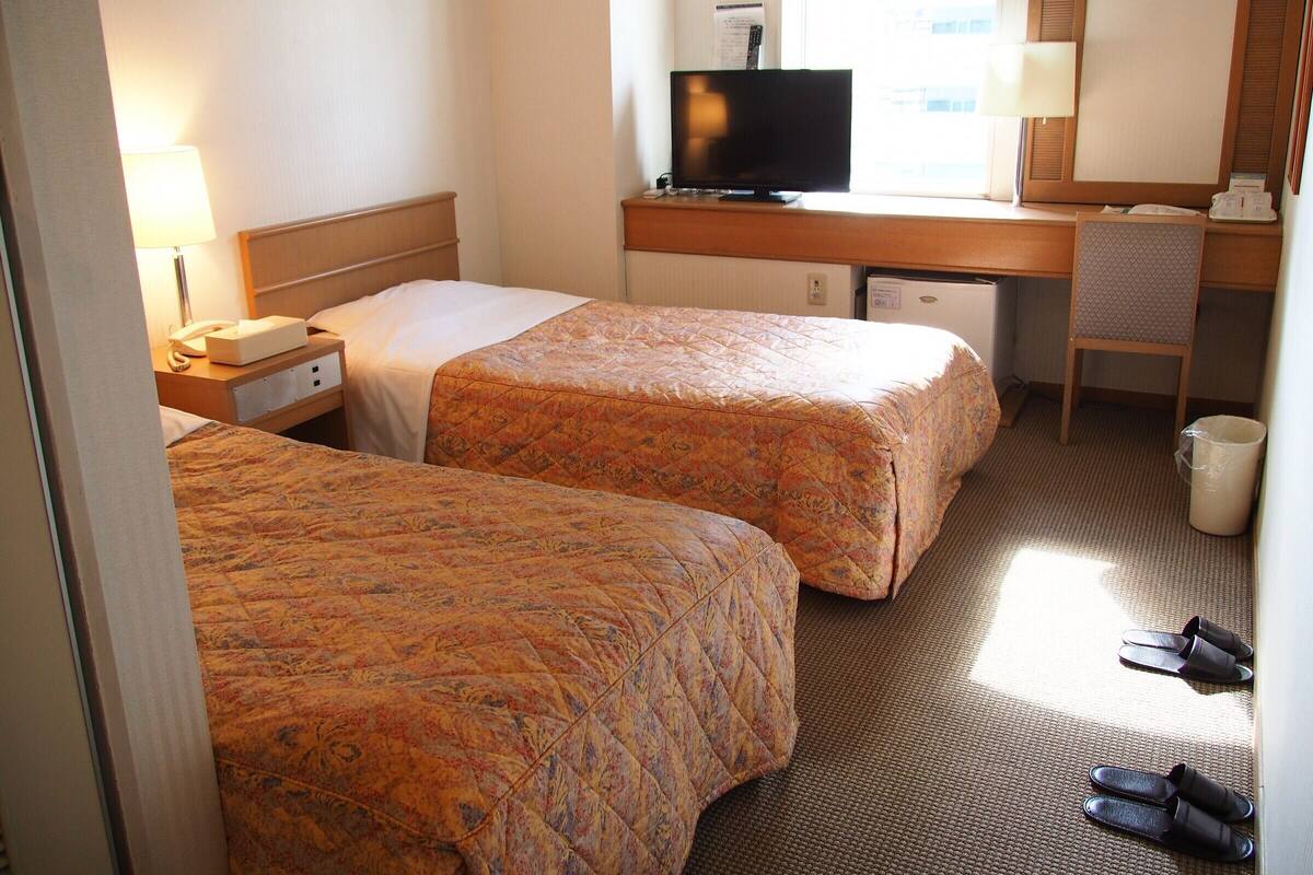 Une chambre d’hôtel avec deux lits, un bureau avec une télévision, une chaise et une poubelle.