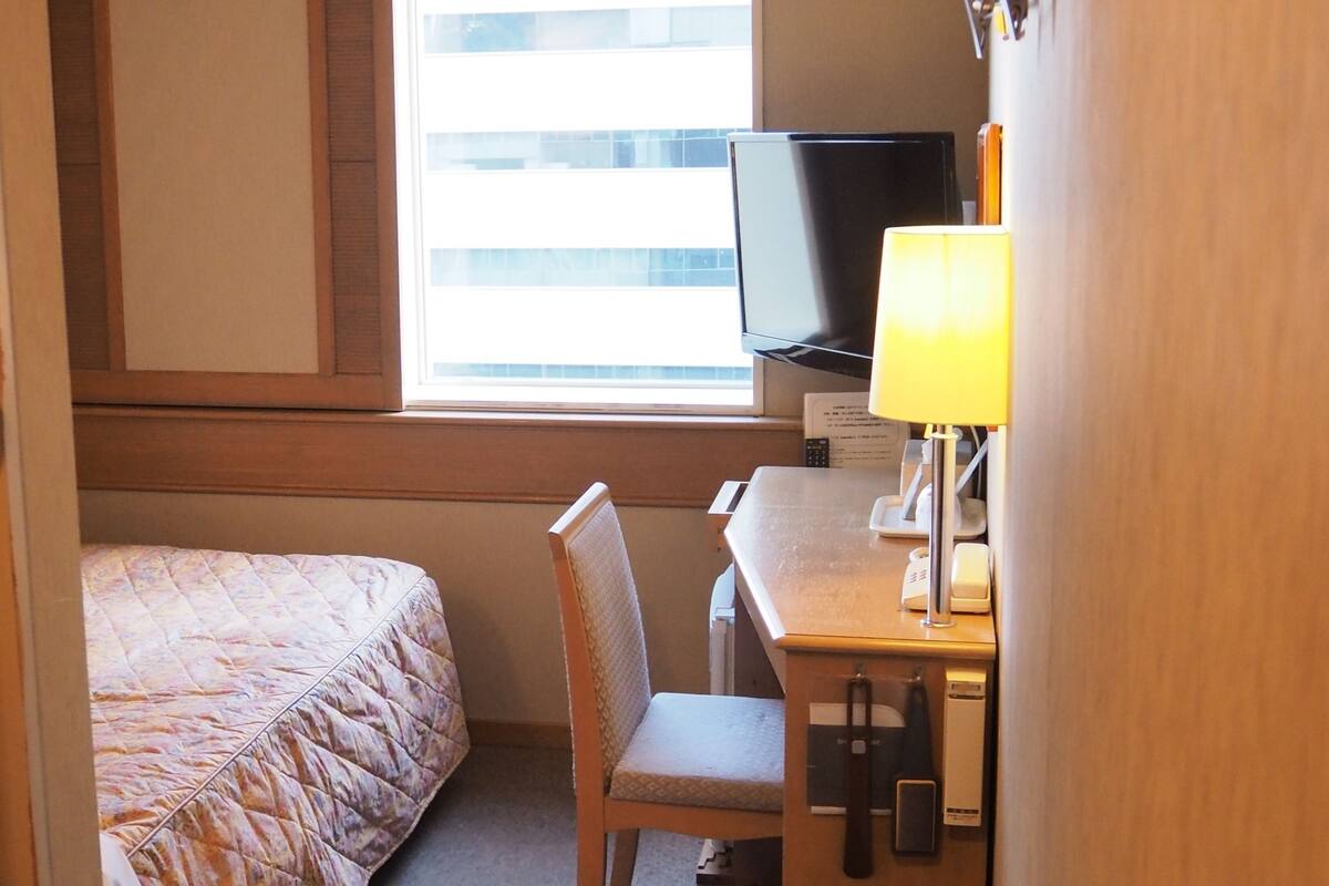 Une chambre d’h ôtel avec un lit, un bureau avec une lampe, une télévision et une fenêtre avec des stores.