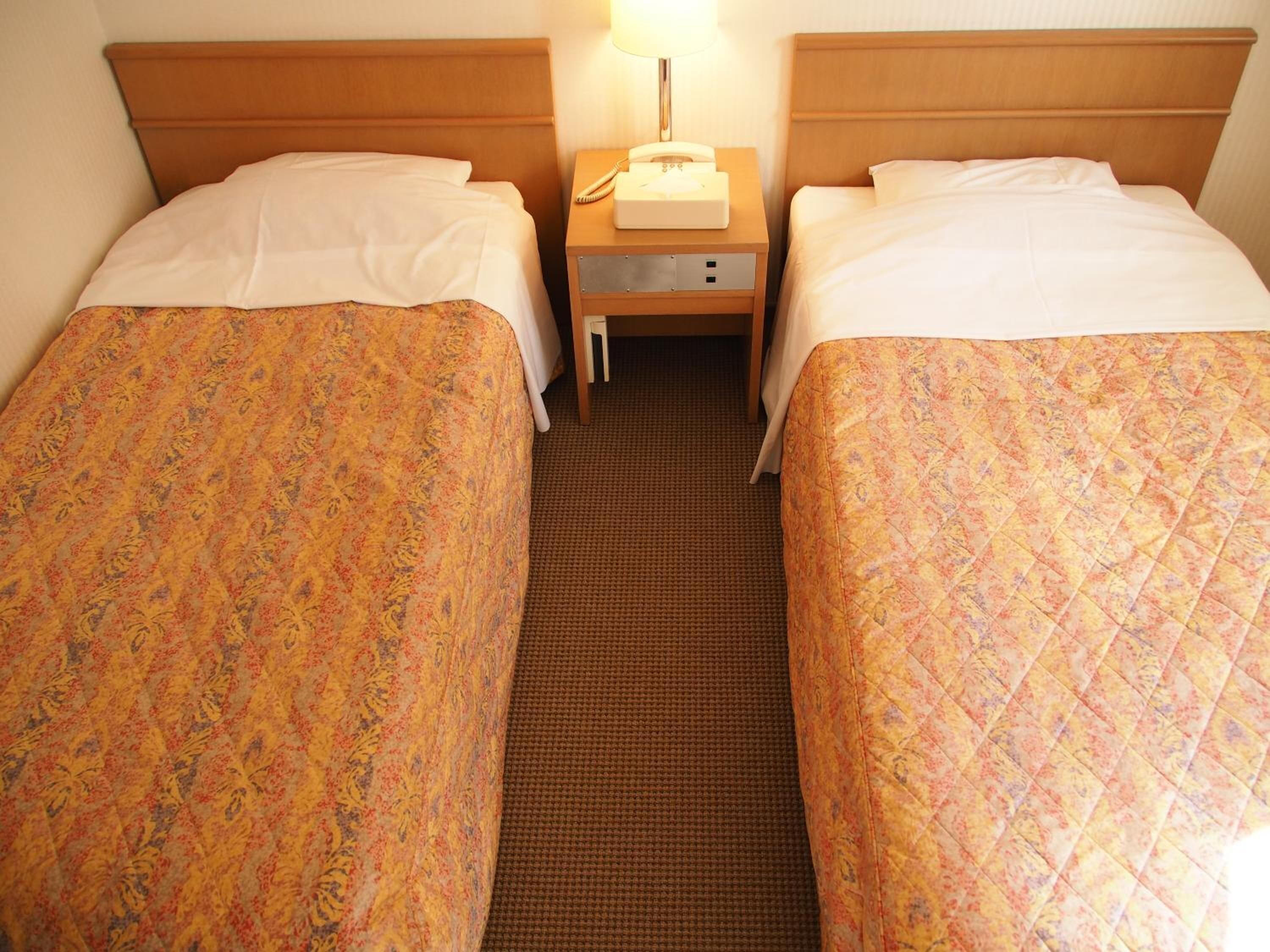 A hotel room with two single beds, each with a floral-patterned bedspread and white pillows. There is a nightstand between the beds with a lamp and a telephone.