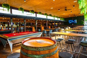 Zona de bar con mesas de billar, taburetes y plantas colgantes.