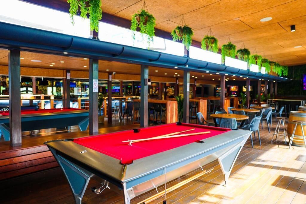 Une table de billard recouverte d’un drap de feutre rouge, entourée de tables et de chaises dans un espace intérieur bien éclairé, agrémenté de plantes suspendues.