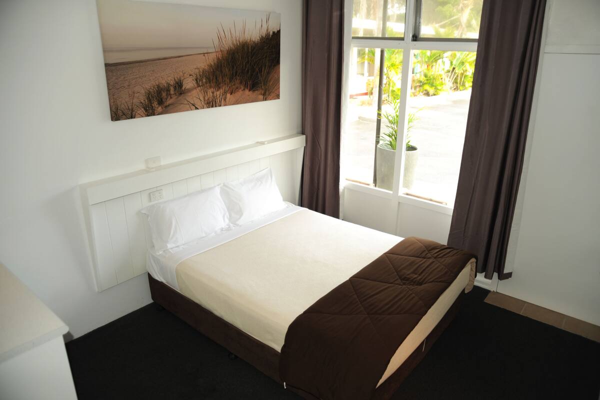 A neatly made bed with white and brown bedding, a large window with curtains, and a painting of a beach scene on the wall.