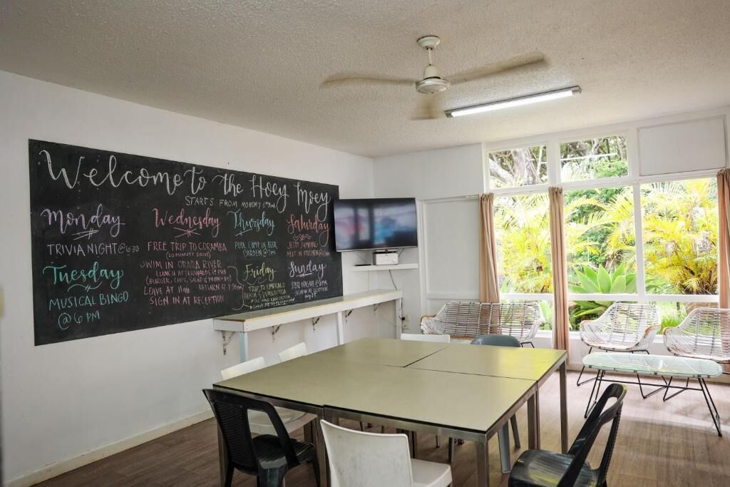 Habitación con pared de pizarra que enumera actividades diarias, un televisor, una mesa con sillas y vistas a zonas verdes a través de amplios ventanales.