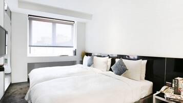 A modern hotel room with a large bed, a bedside table with a phone and books, and a window with blinds.
