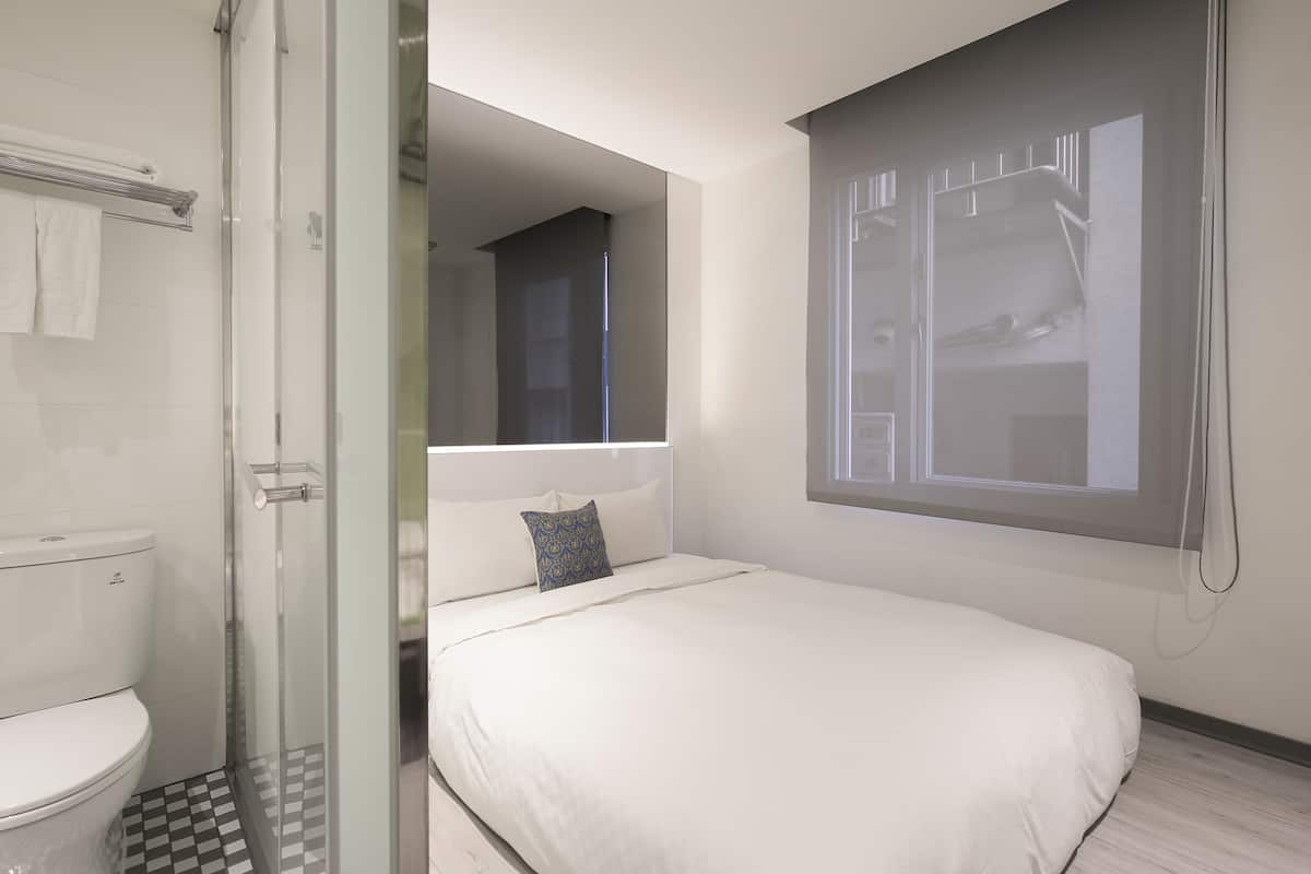A modern hotel room with a glass shower enclosure, a white bed with a blue pillow, and a large window with blinds.