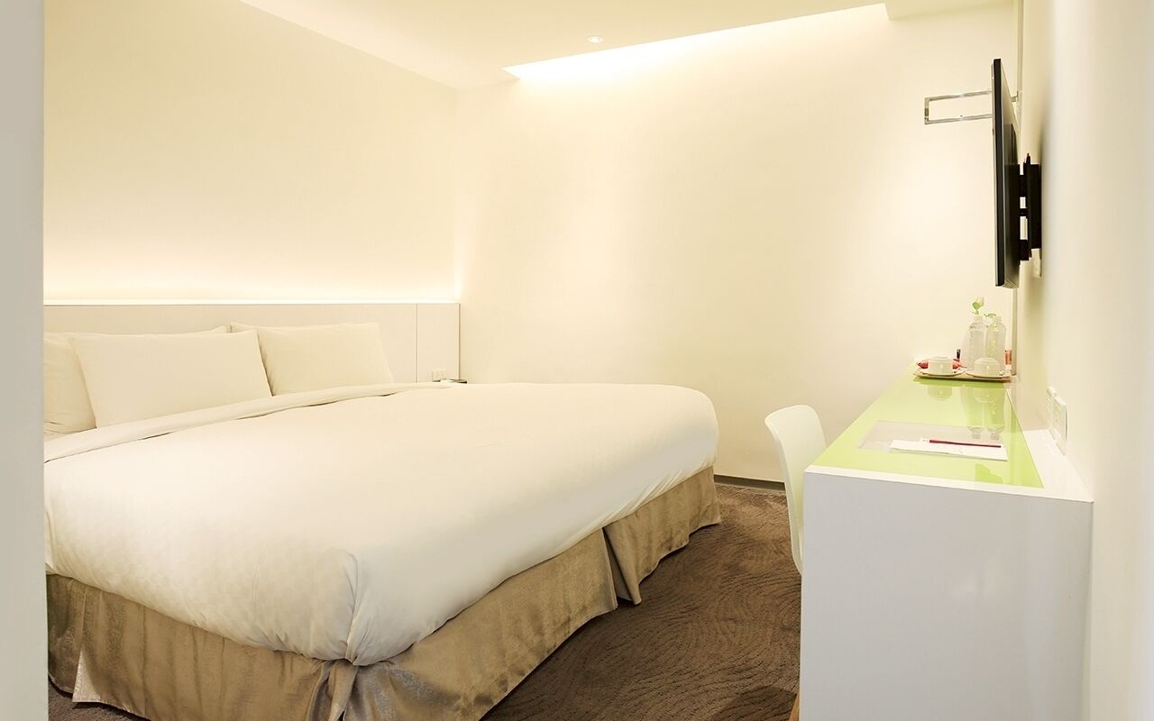 A hotel room with a bed, a desk, a chair, a flat-screen TV mounted on the wall, and a small table with a glass and a bottle.