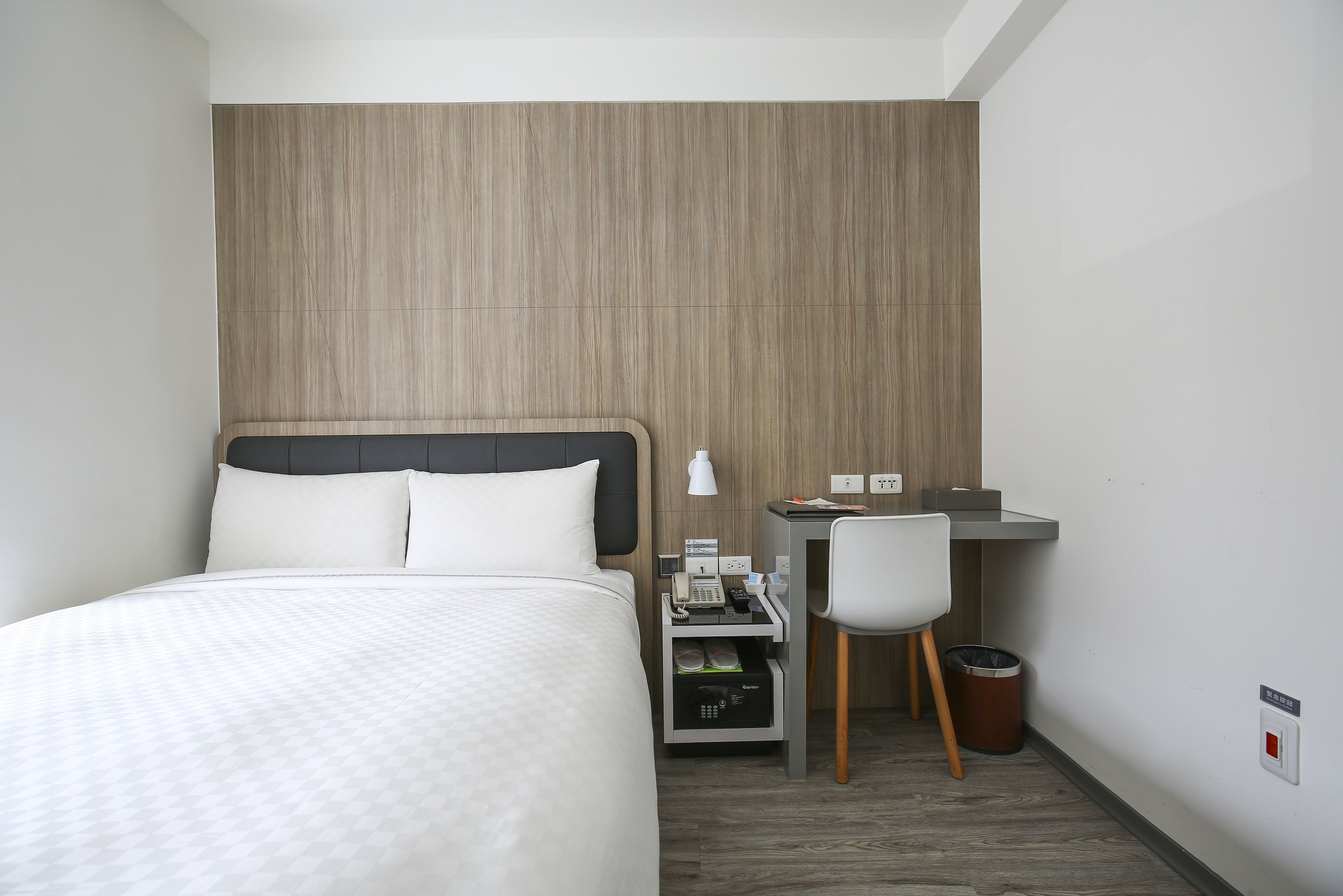 A hotel room with a bed, a desk, a chair, a trash bin, and a wall-mounted phone.