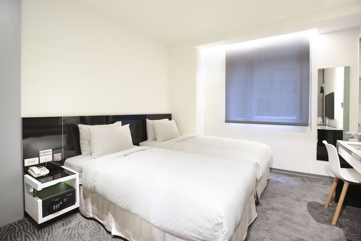 A hotel room with a large bed, a desk, a chair, a nightstand with a phone, and a window with blinds.