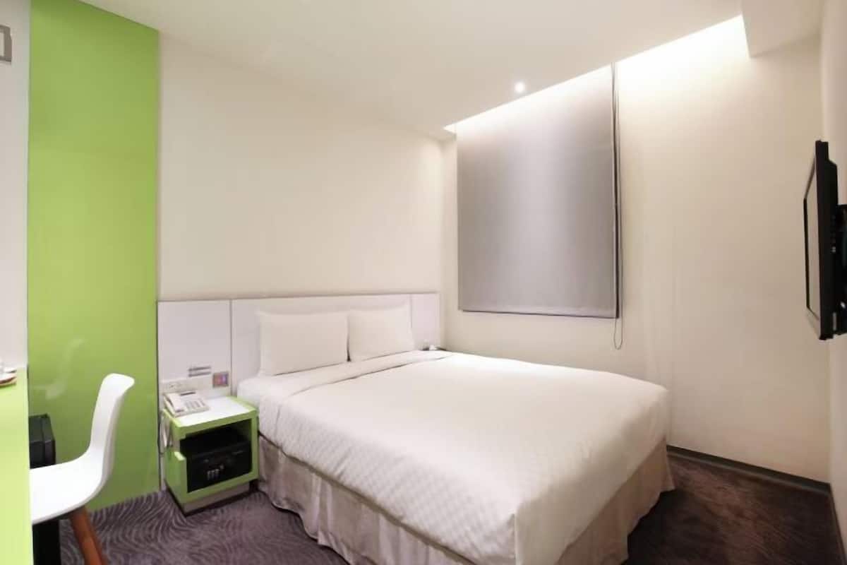 A hotel room with a bed, a bedside table with a phone, a chair, and a TV mounted on the wall.