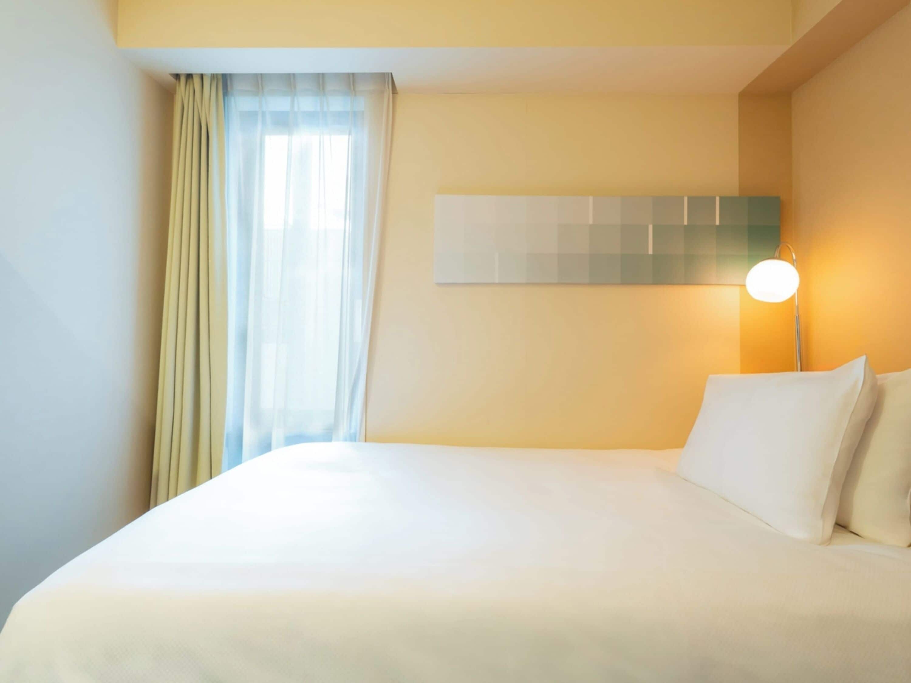 A hotel room with a bed, a window with curtains, a wall-mounted light, and a plain wall.
