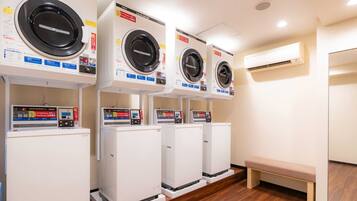 A row of white front-loading washing machines with control panels, a wooden bench, and an air conditioning unit on the wall.
