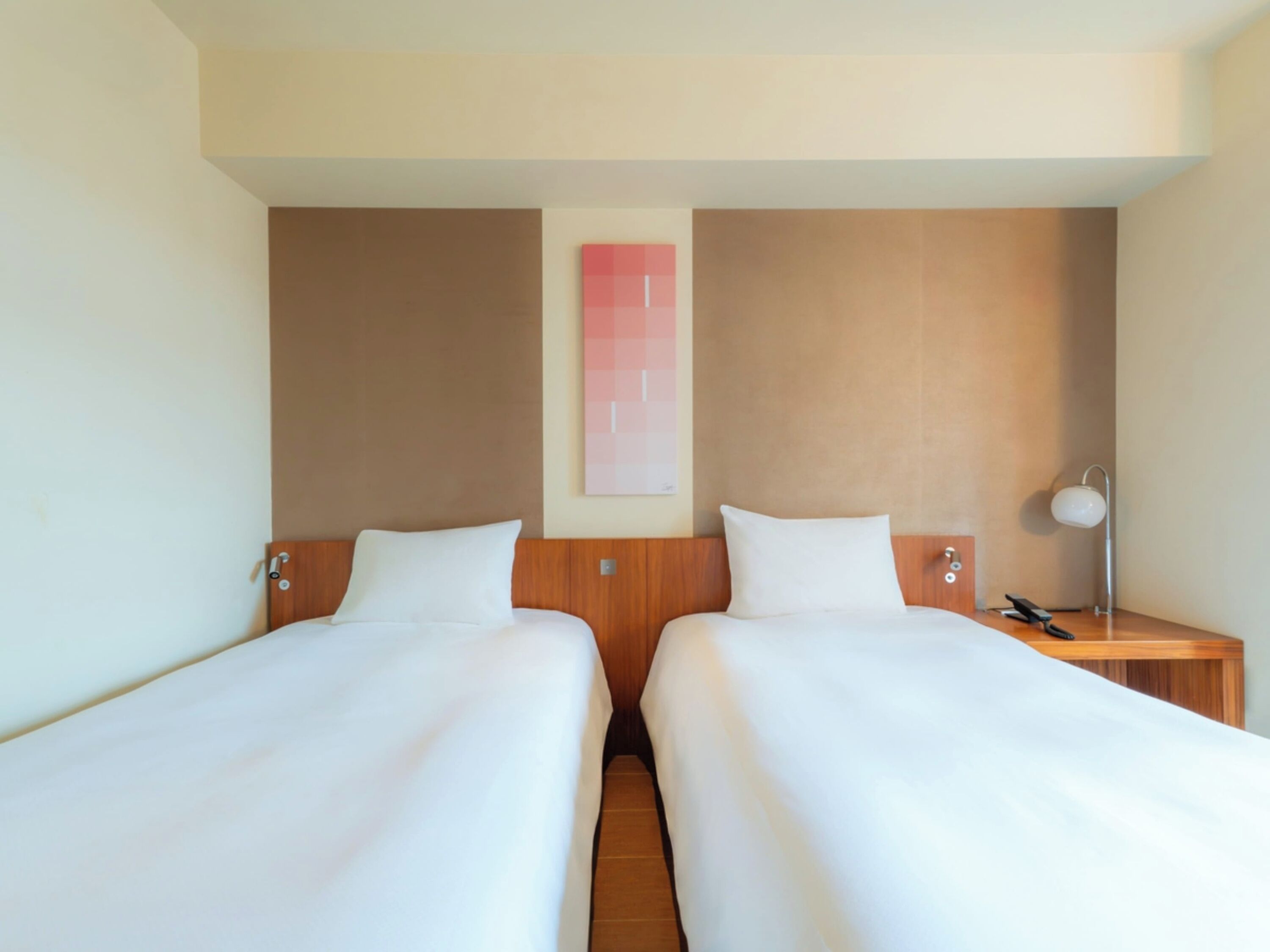 Habitació d'hotel amb dos llits individuals, un panell de fusta al capçal, una taula de nit amb una làmpada i una obra d'art penjada a la paret.