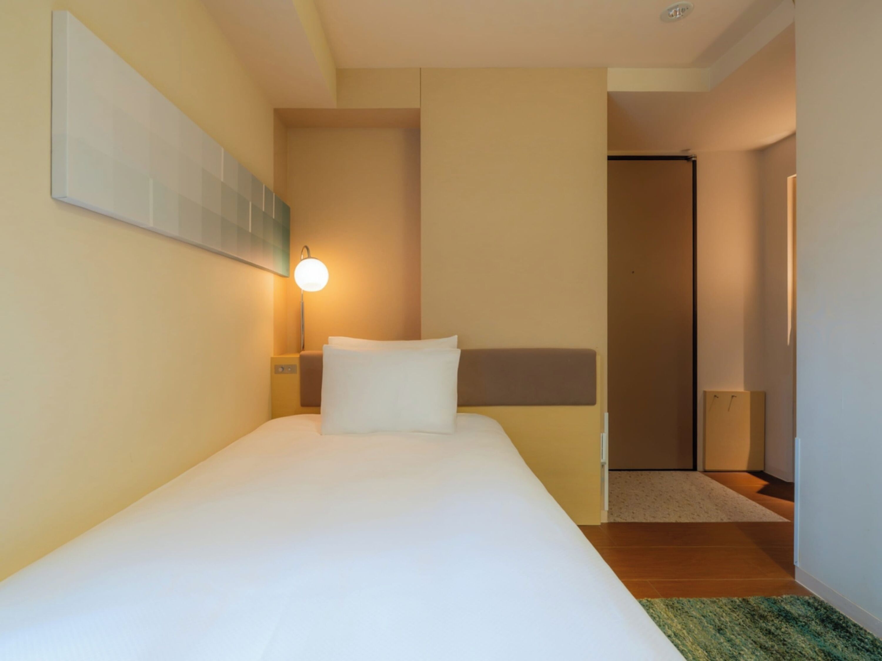 A hotel room with a bed, a nightstand, a wall-mounted lamp, and a door leading to another room.