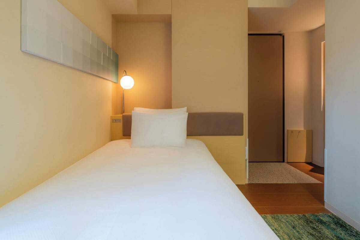A hotel room with a bed, a nightstand, a wall-mounted lamp, and a door leading to another room.