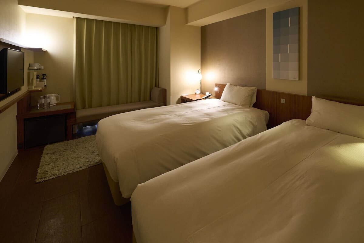 Camera d'albergo con due letti, una scrivania, una sedia, una TV e una finestra con tende.