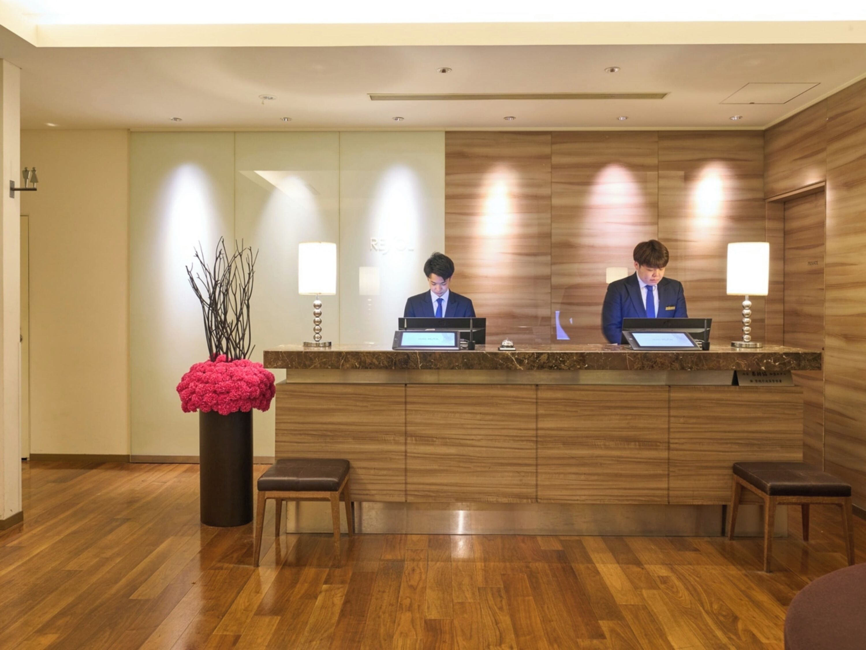 A reception desk with two staff members, a wooden floor, and a vase with branches.