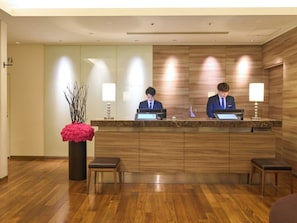 A reception desk with two staff members, a wooden floor, and a vase with branches. - Hotel Resol Ikebukuro (Tokyo)