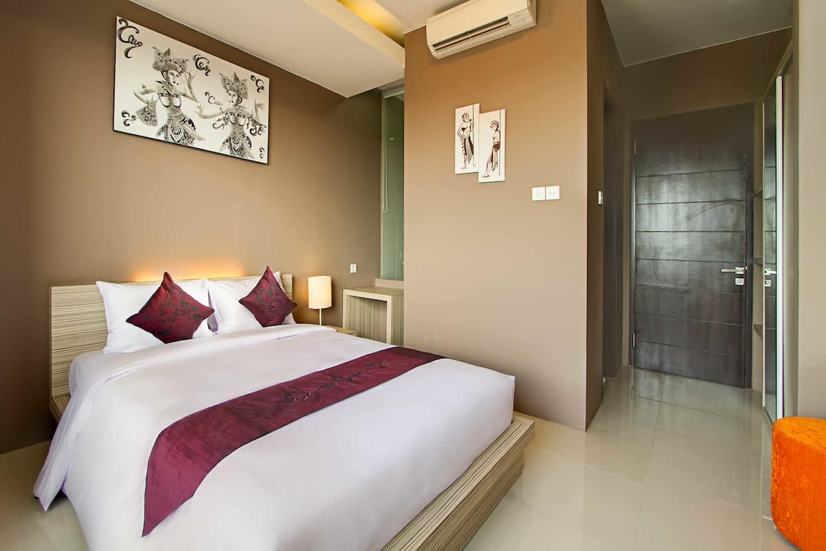 A hotel room with a large bed, a nightstand, a wall-mounted air conditioner, and a door leading to another room.