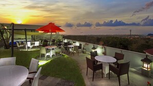 An outdoor patio with tables and chairs, a red umbrella, and a view of the sunset and city skyline. - Parkside DenBukit Residence & Suite Jimbaran (Jimbaran)