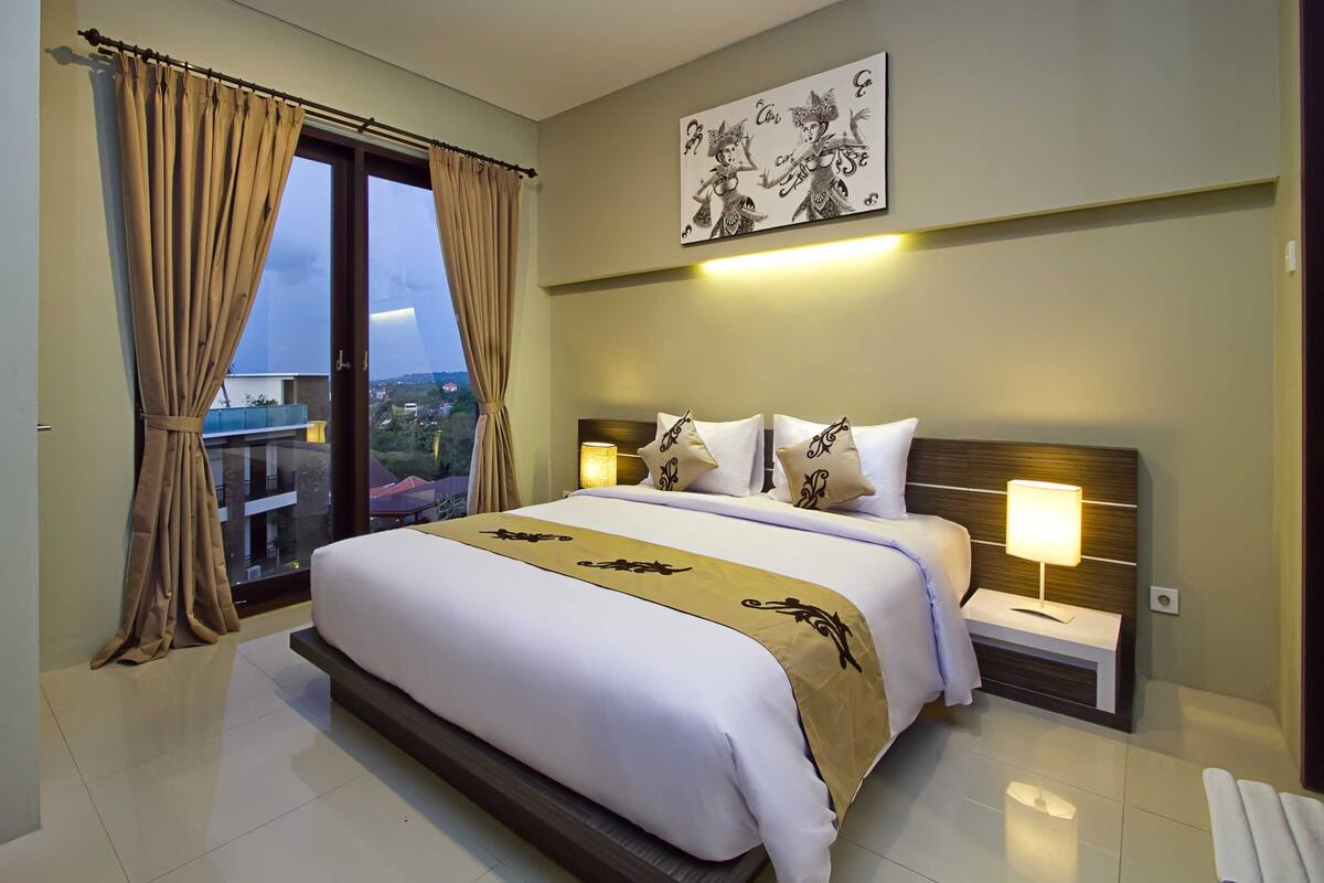 A hotel room with a large bed, a nightstand with a lamp, and a view of the city through the window.