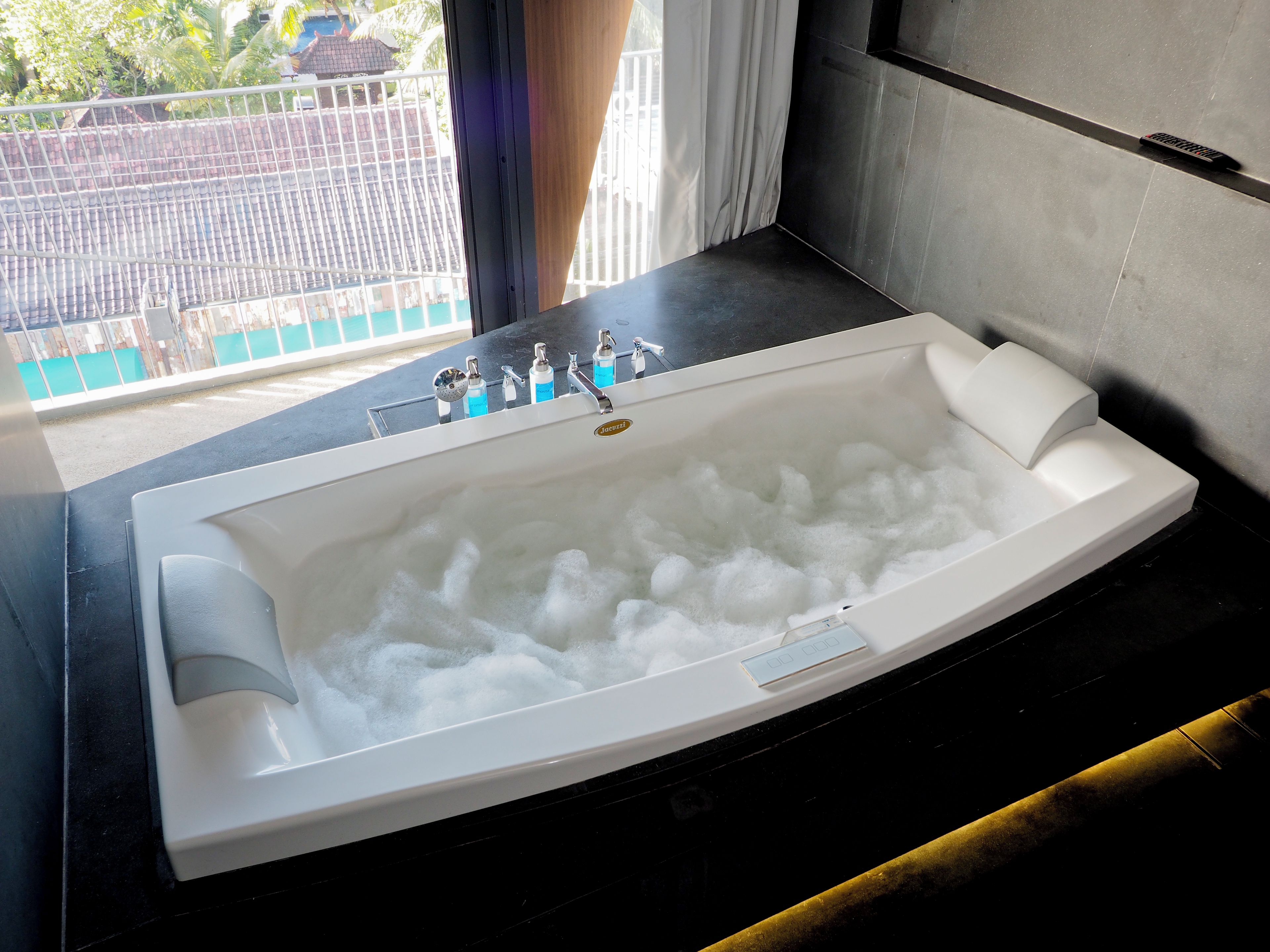 A large bathtub with multiple faucets and a control panel, situated in a modern bathroom with a view of a balcony.