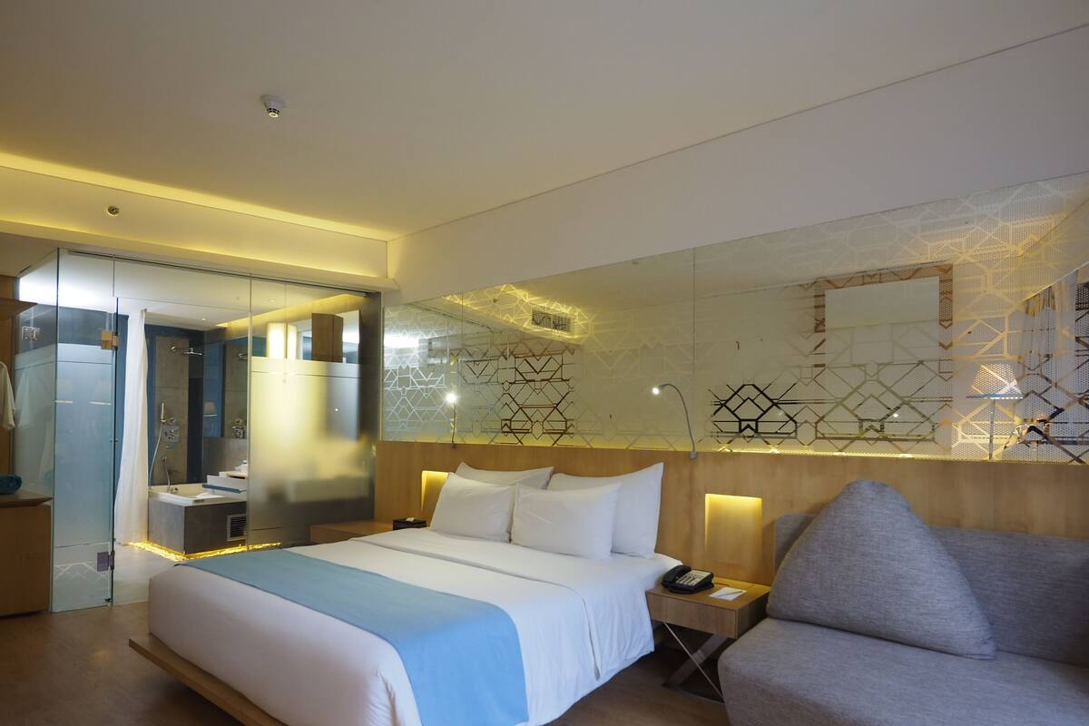 A modern hotel room with a large bed, a bedside table with a lamp, a sofa, and a glass-enclosed shower area.