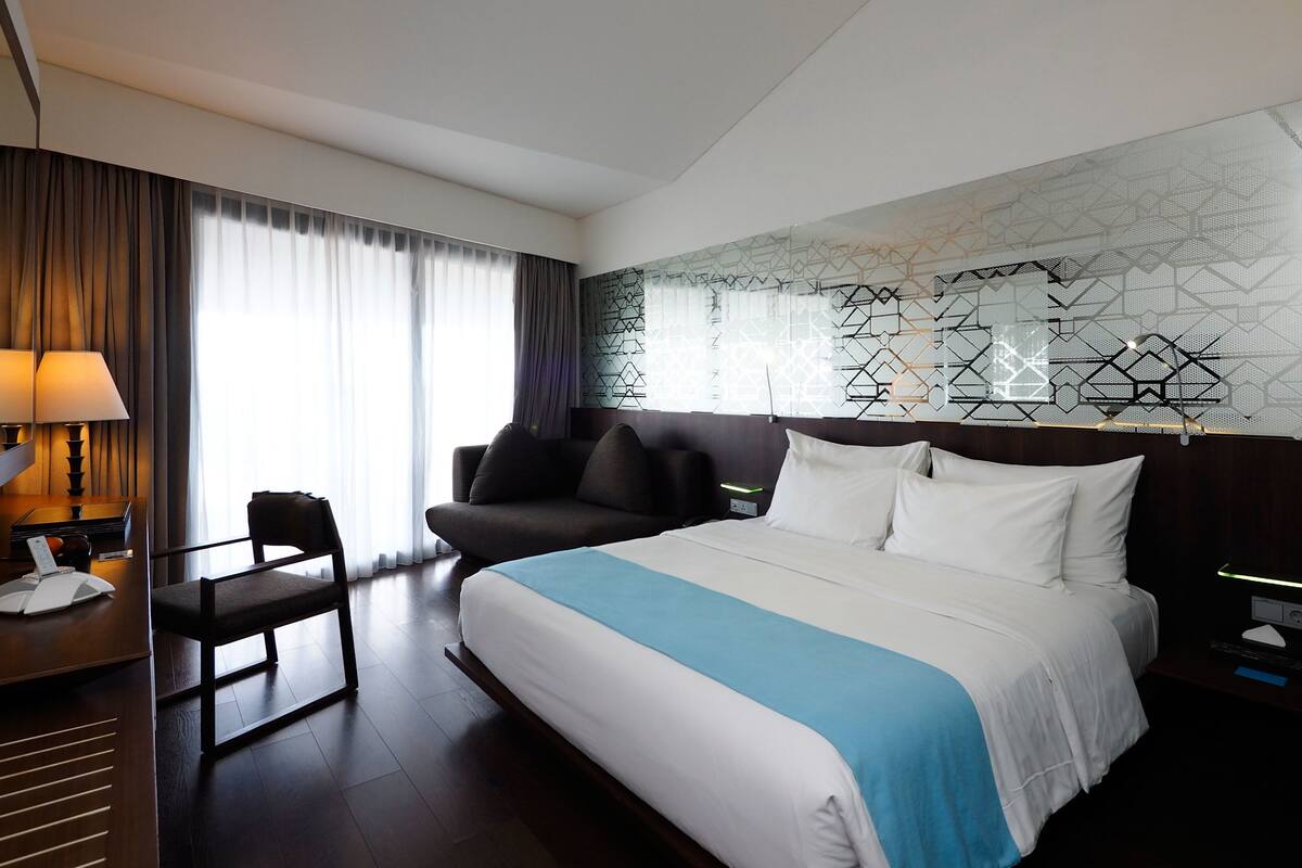A hotel room with a large bed, a desk with a lamp, a chair, and a window with curtains.