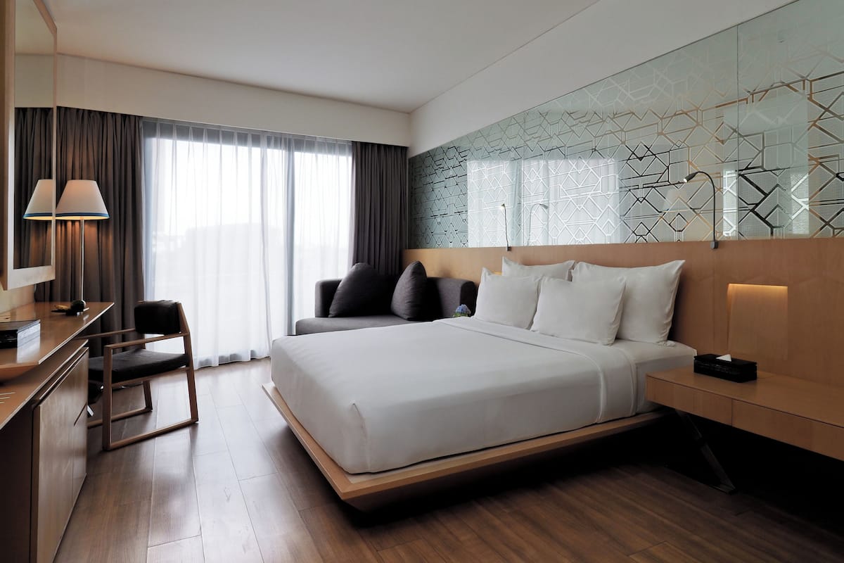 A modern hotel room with a large bed, a desk, a chair, and a large window with curtains.