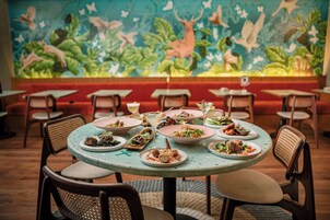 Um restaurante com mesas e cadeiras redondas, um mural na parede e diversos pratos sobre as mesas.
