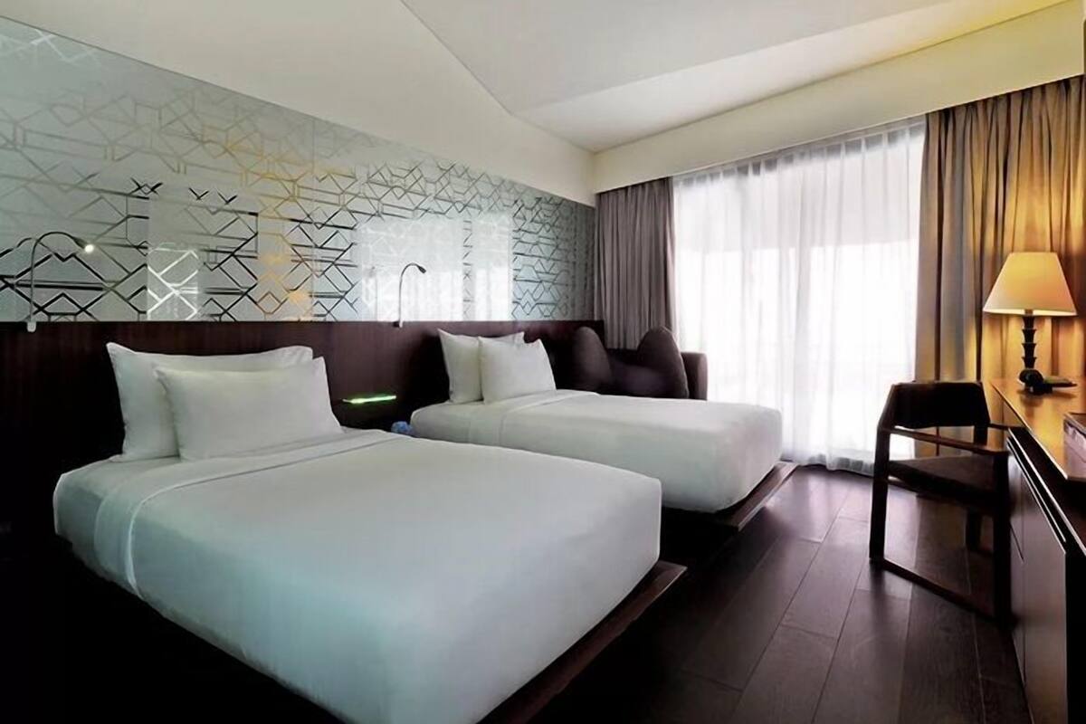 A hotel room with two beds, a desk with a chair, a lamp, and a window with curtains.
