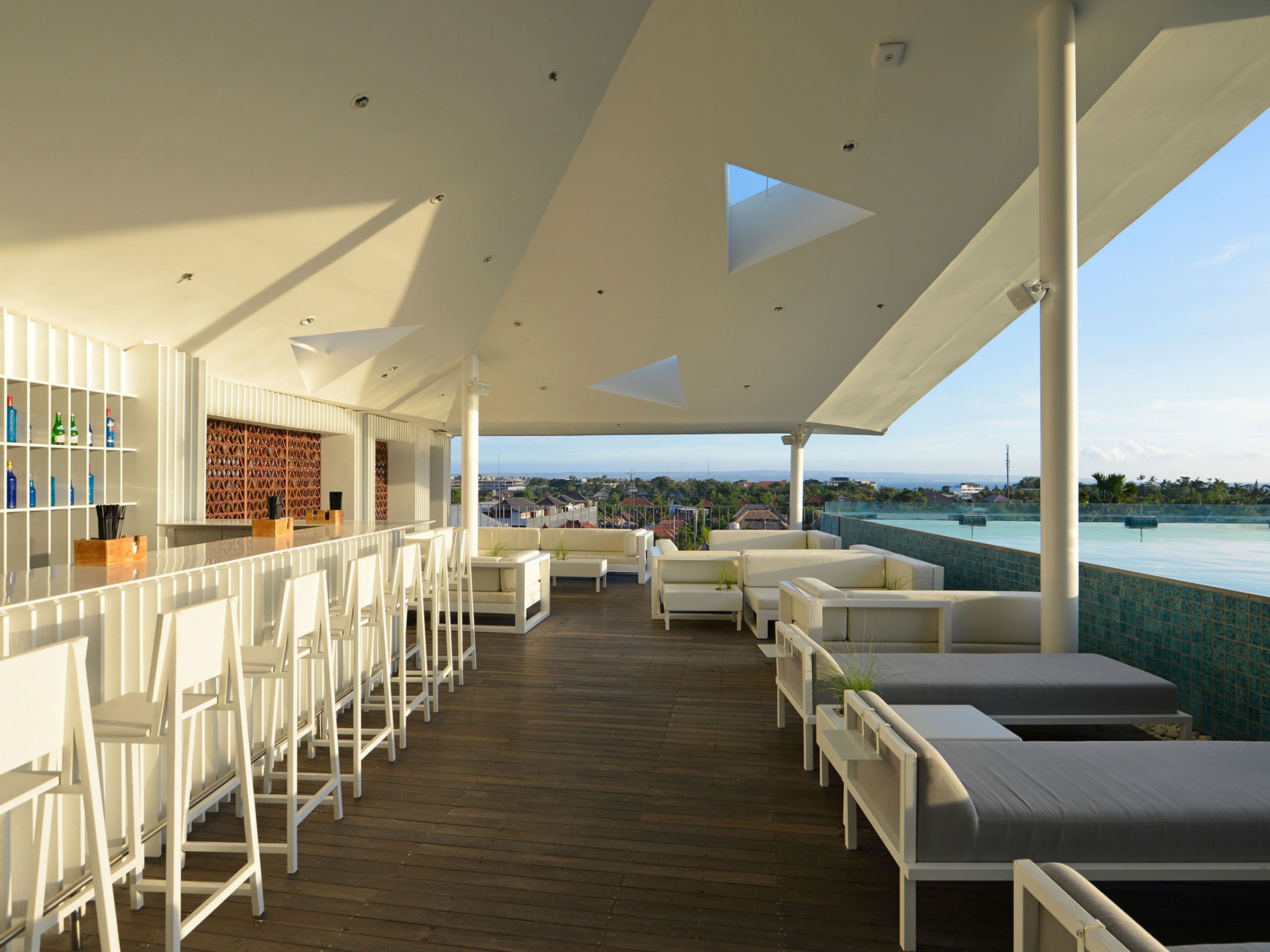 Un espace de détente extérieur moderne, avec des meubles blancs, un bar doté d’étagères remplies de bouteilles et une vue sur le paysage urbain.