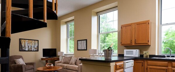 A spacious living area with a sofa, a television, and artwork on the walls. There is a kitchen with wooden cabinets and a microwave.