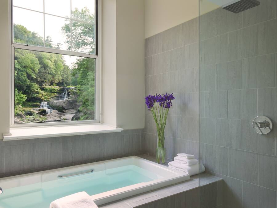 A bathroom with a bathtub, a window overlooking a waterfall, and a vase with purple flowers.