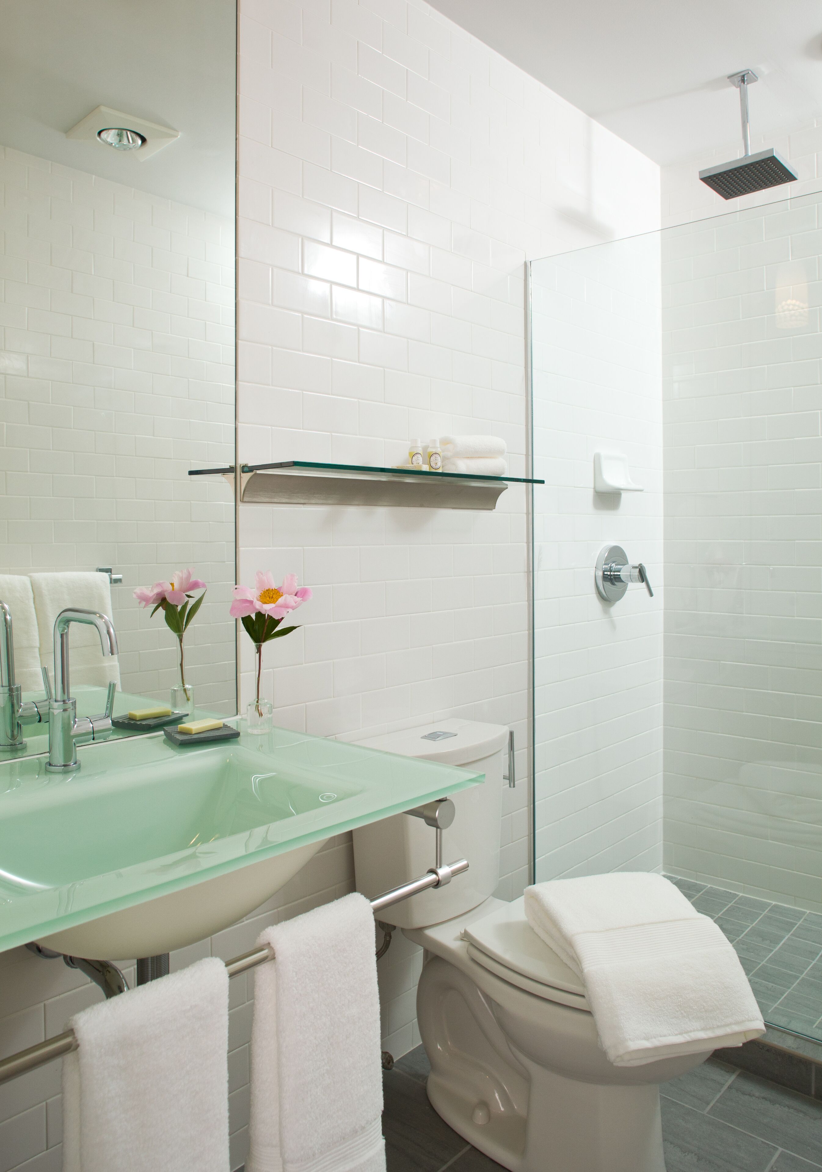 A modern bathroom with a glass sink, white tiles, a glass shower enclosure, and a toilet with a towel on it.