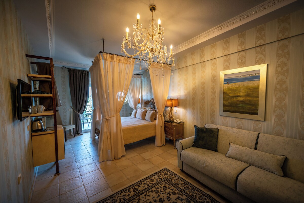 A bedroom with a bed, a sofa, a chandelier, a nightstand, and a painting on the wall.