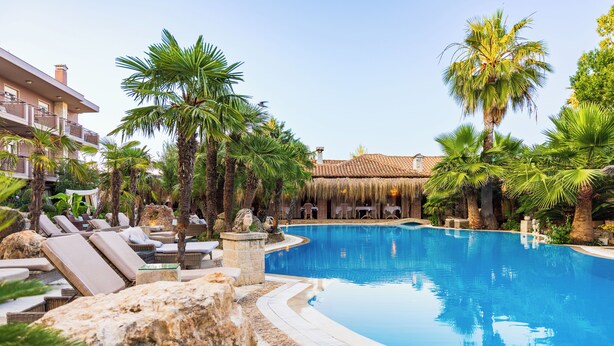 A pool surrounded by lounge chairs and palm trees, with a building in the background. - Achtis Boutique Hotel (Kassandra)