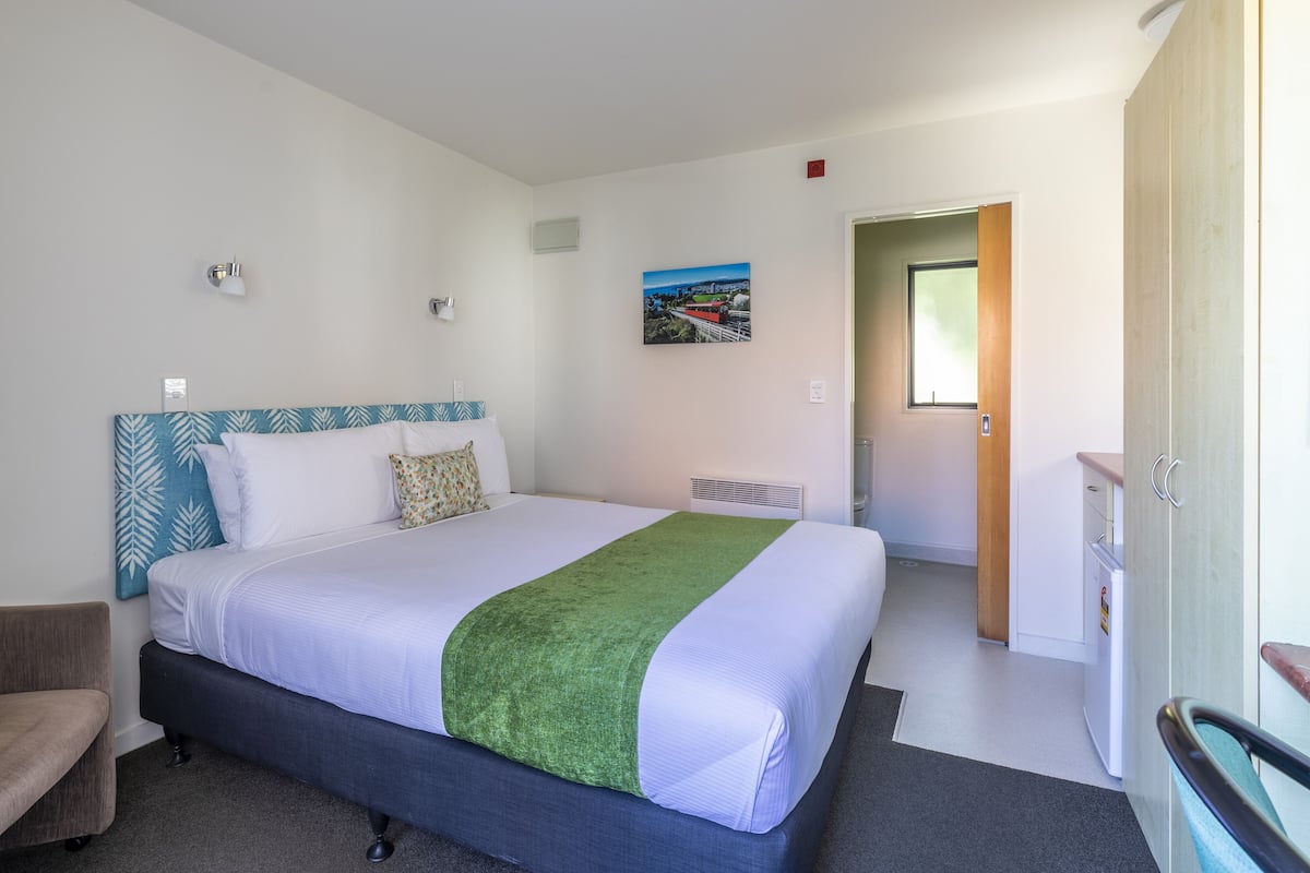 Ein Hotelzimmer mit einem großen Bett, einem kleinen Schreibtisch, einem Stuhl, einem Bild an der Wand und einer Tür zu einem anderen Raum.