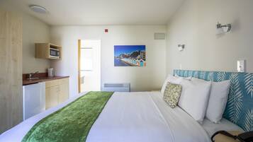 A hotel room with a bed, a green throw blanket, a microwave, a small kitchenette, and a painting of a coastal scene.