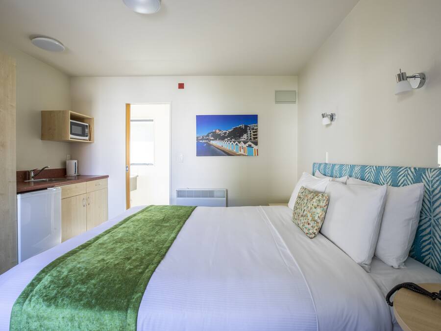 A hotel room with a bed, a green throw blanket, a microwave, a small kitchenette, and a painting of a coastal scene.