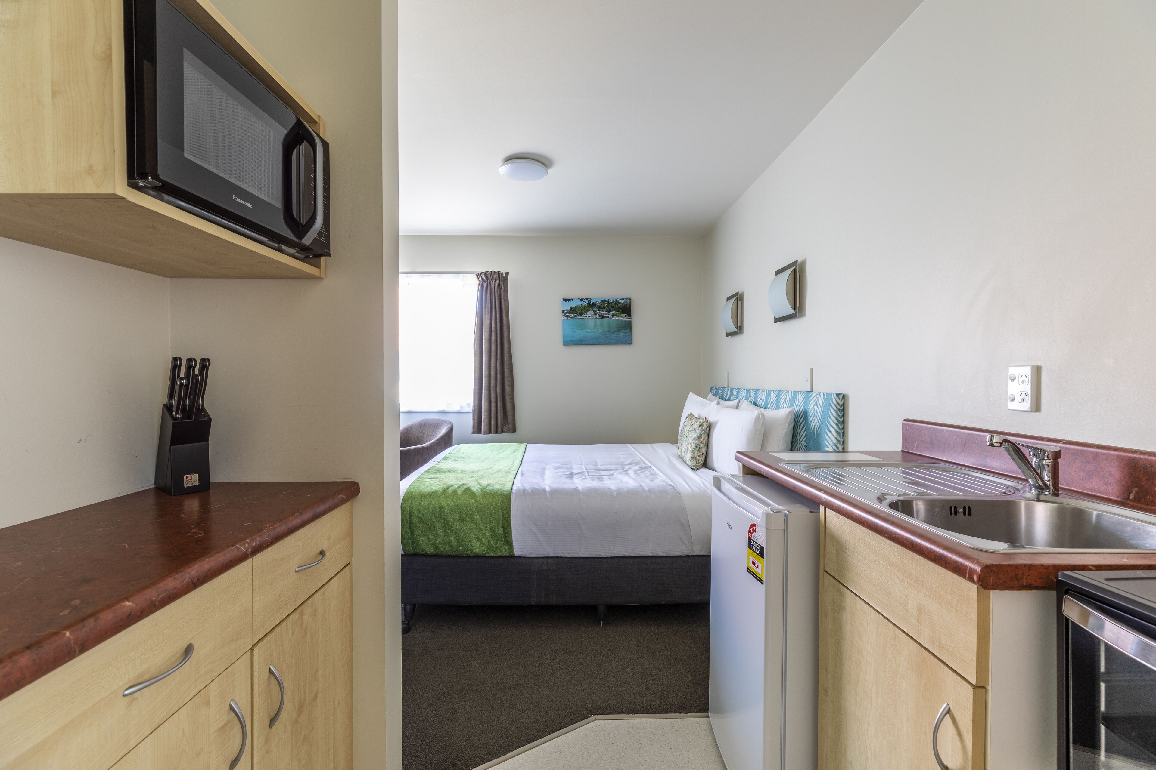 Une chambre d’hôtel compacte comprenant un coin cuisine, un lit avec une couverture verte, un petit coin salon et une fenêtre avec des rideaux.