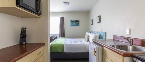 Une chambre d’hôtel compacte comprenant un coin cuisine, un lit avec une couverture verte, un petit coin salon et une fenêtre avec des rideaux.