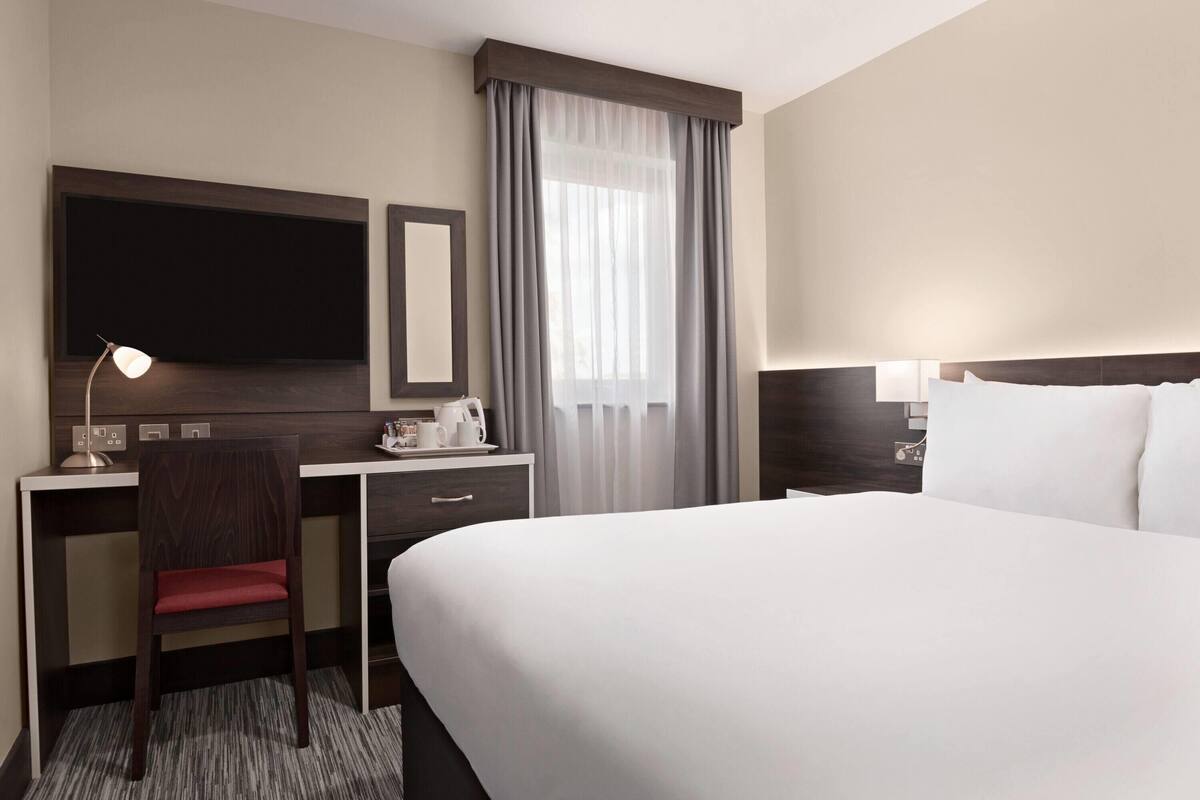 A hotel room with a bed, desk, chair, TV, and window with curtains.